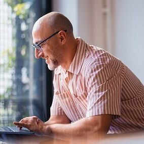 A cheerful man working on his laptop at a desk in a modern home interior, illuminated by natural light and surrounded by positive vibes, showcasing productivity, comfort, and a relaxed atmosphere.